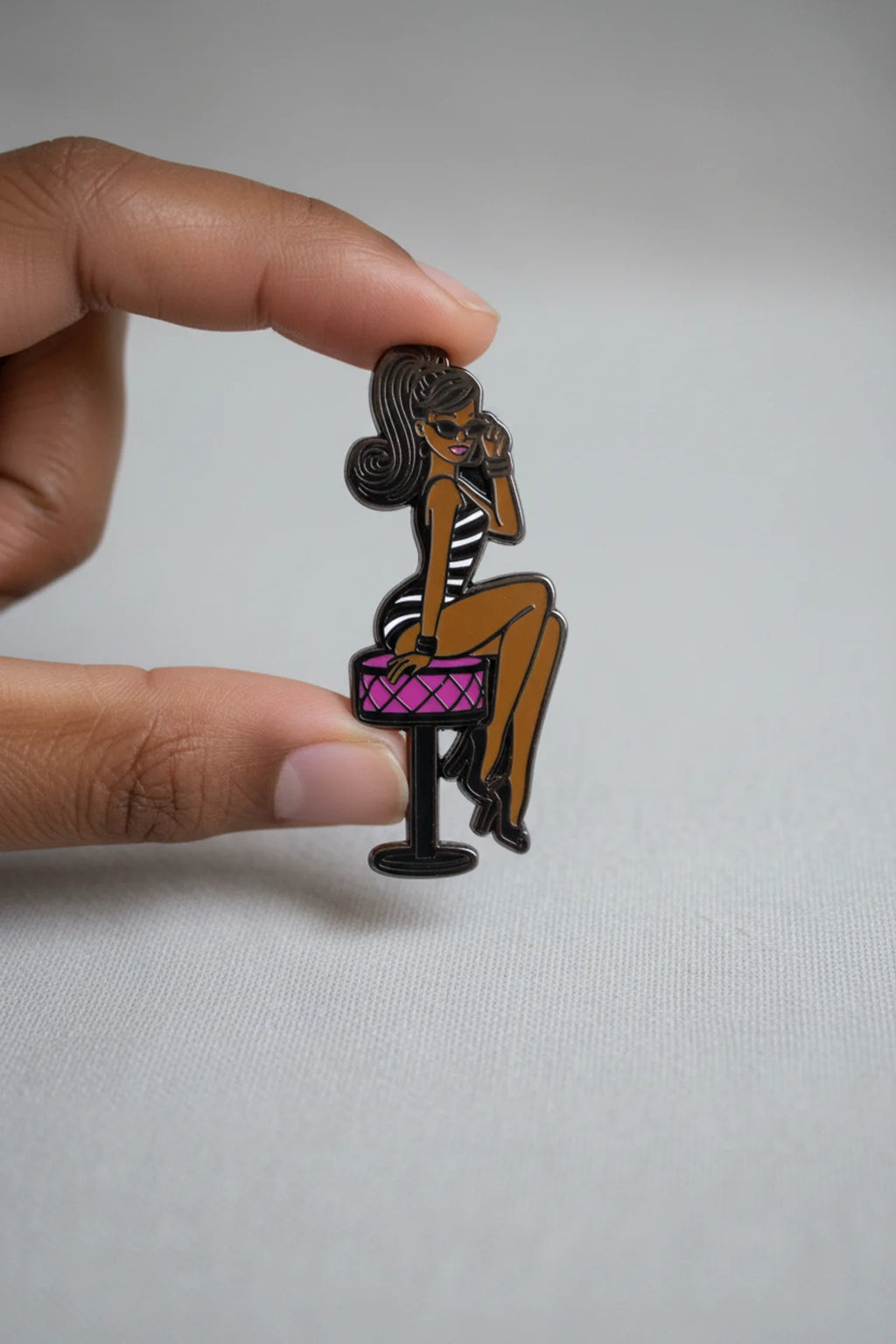 Pin of a woman sitting on a stool with a pink seat held by a hand against a gray background
