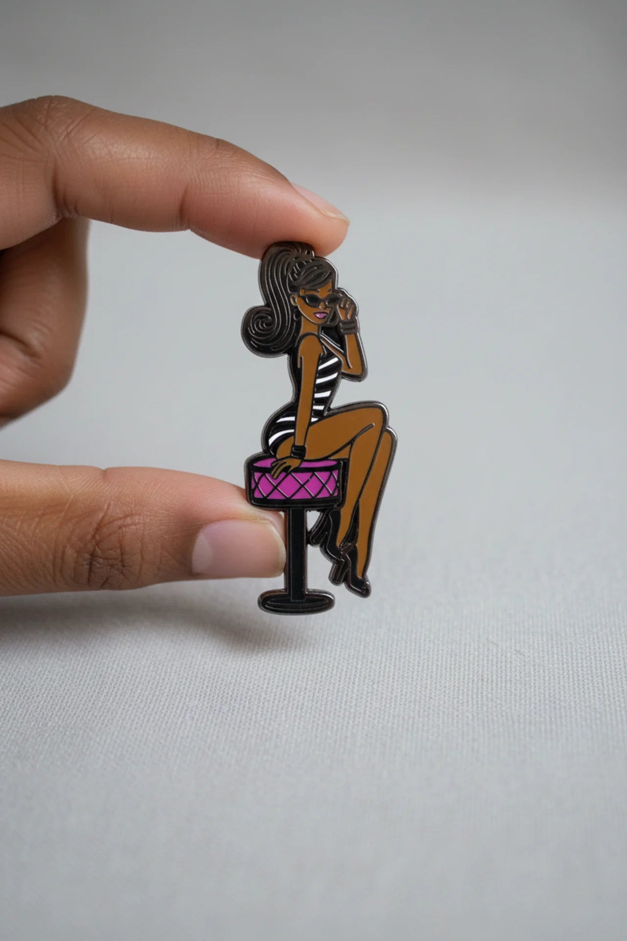 Pin of a woman sitting on a stool with a pink seat held by a hand against a gray background